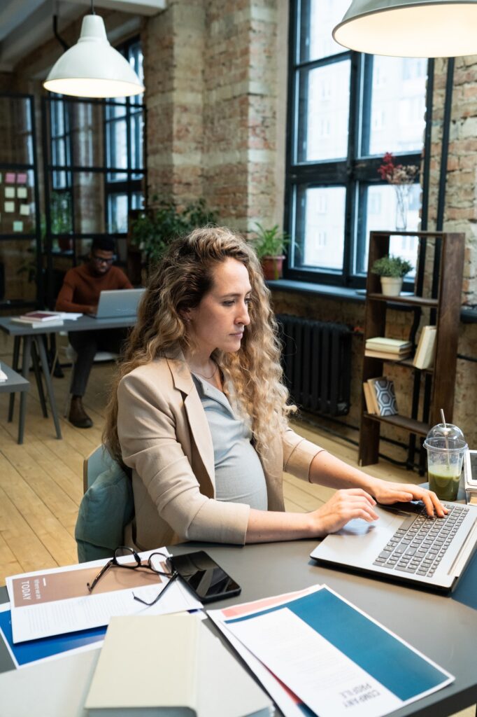 Young serious pregnant accountant sitting by desk in front of laptop